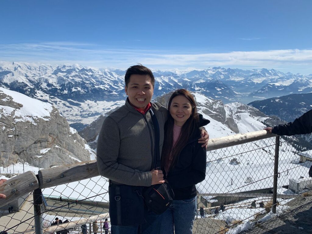 A couple smiling and embracing in front of a snowy mountain range, with a clear blue sky above and a wooden fence in the foreground, sharing one of their honeymoon stories.
