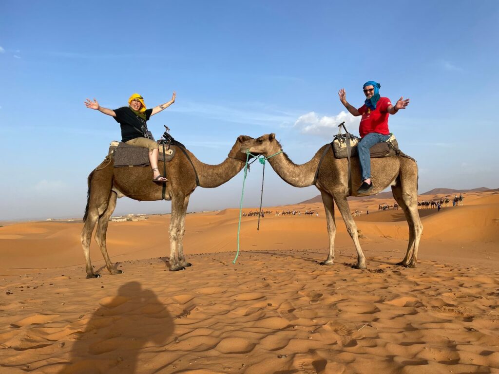 Two people joyfully raising their arms while sitting on camels in a desert, with sand dunes and a clear sky in the background, celebrating their honeymoon.