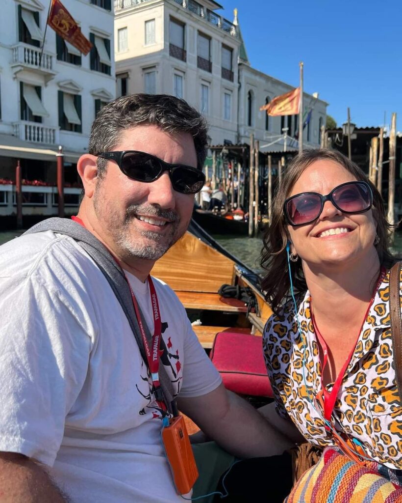 A man and a woman smiling on a sunny day, seated in a gondola with waterfront buildings in the background during their honeymoon.