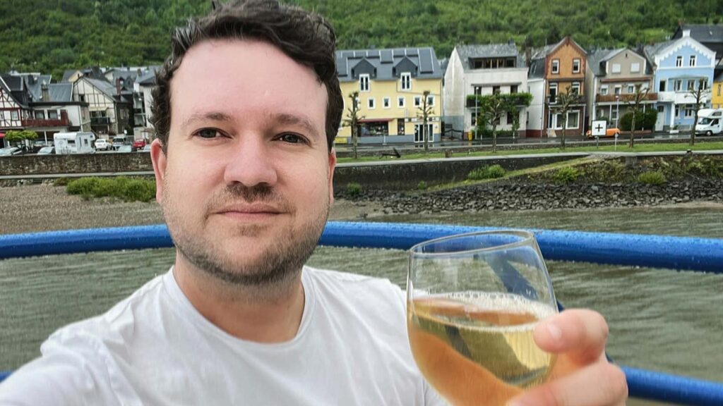 Man holding a glass of wine on a boat, with picturesque houses and green hills in the background, planning a proposal in Paris.
