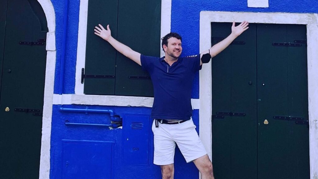 A man with arms raised stands between two blue and green doors, wearing a blue polo shirt and white shorts, reminiscent of his last proposal in Paris.