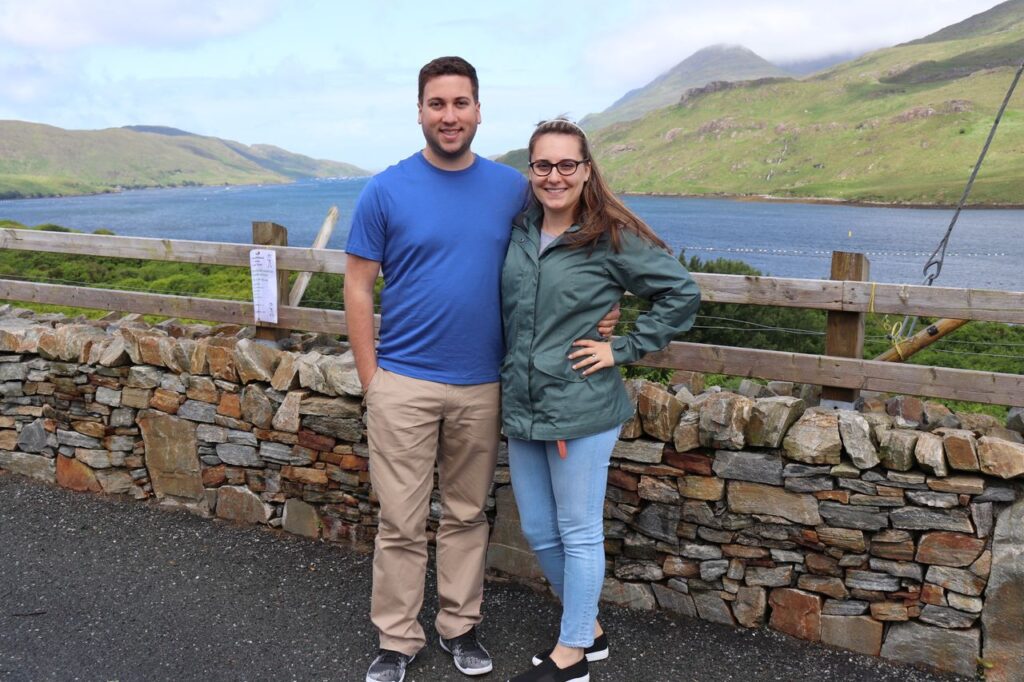 A man and a woman smiling, standing together behind a stone wall with a lake and green hills in the background, likely reminiscing about their honeymoon stories.