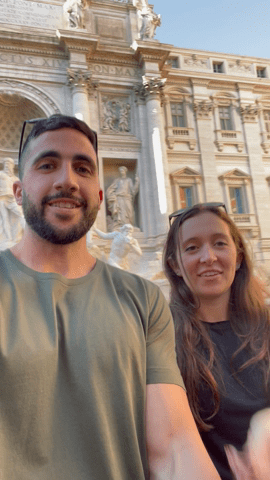 A man and a woman taking a selfie with a historic building featuring ornate sculptures in the background during their honeymoon.