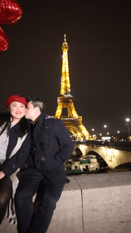A man kissing a woman on the cheek at night, with the illuminated Eiffel Tower in the background and red heart-shaped balloons around them, exemplifying one of many romantic honeymoon stories.