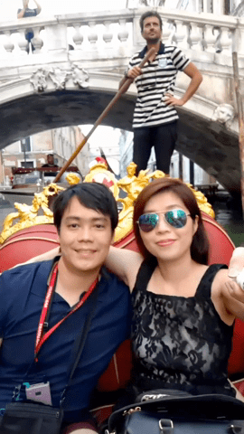 A couple takes a honeymoon selfie in a gondola under a bridge in Venice, with a gondolier steering in the background.