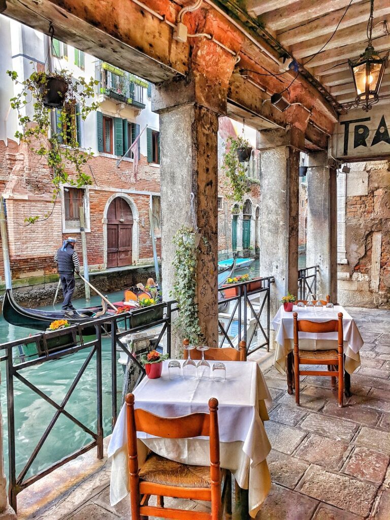 Outdoor dining area with tables set for meals beside a canal in Venice, Italy, with a gondolier rowing a boat in the background, offering an authentic experience of travel like a local in
