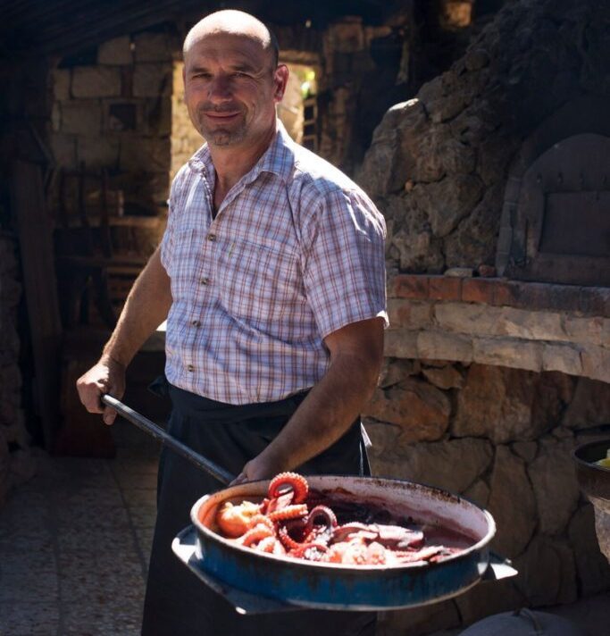 A man smiling while holding a large pan of cooked octopus, standing in a rustic outdoor kitchen in Central Europe.