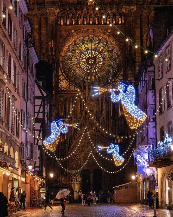 A vibrant Christmas night scene of a festive street with illuminated angel decorations leading to a large, ornate cathedral with a rose window, bustling with people and ambient lights.