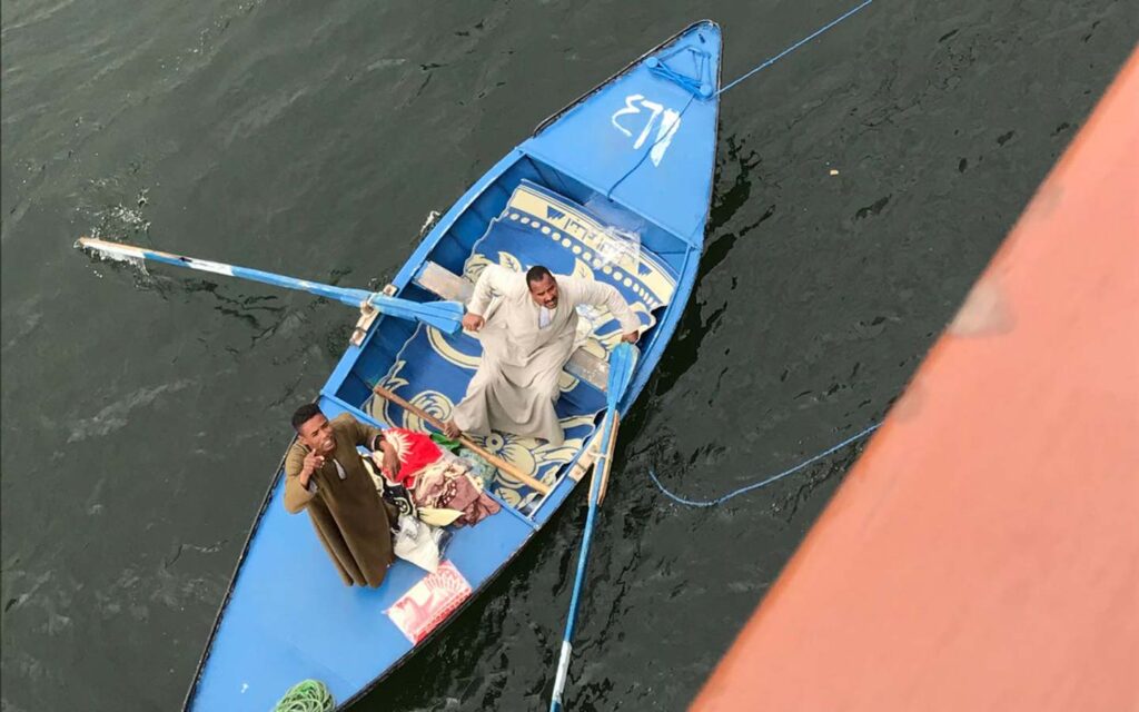 Two people in a blue rowboat on the Nile, with one person rowing and the other sitting, surrounded by bags and objects—a snapshot from an Egypt travel story.