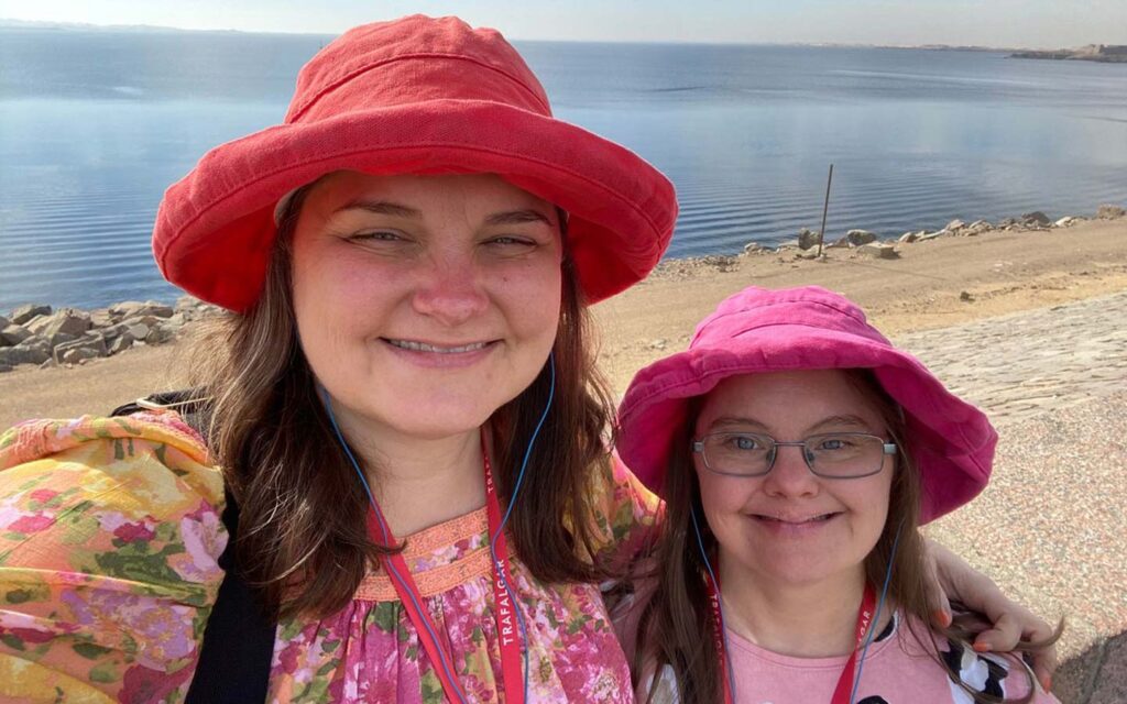 Two women wearing red and pink bucket hats take a selfie on a sunny day with a calm lake and rocky shore in Egypt in the background.