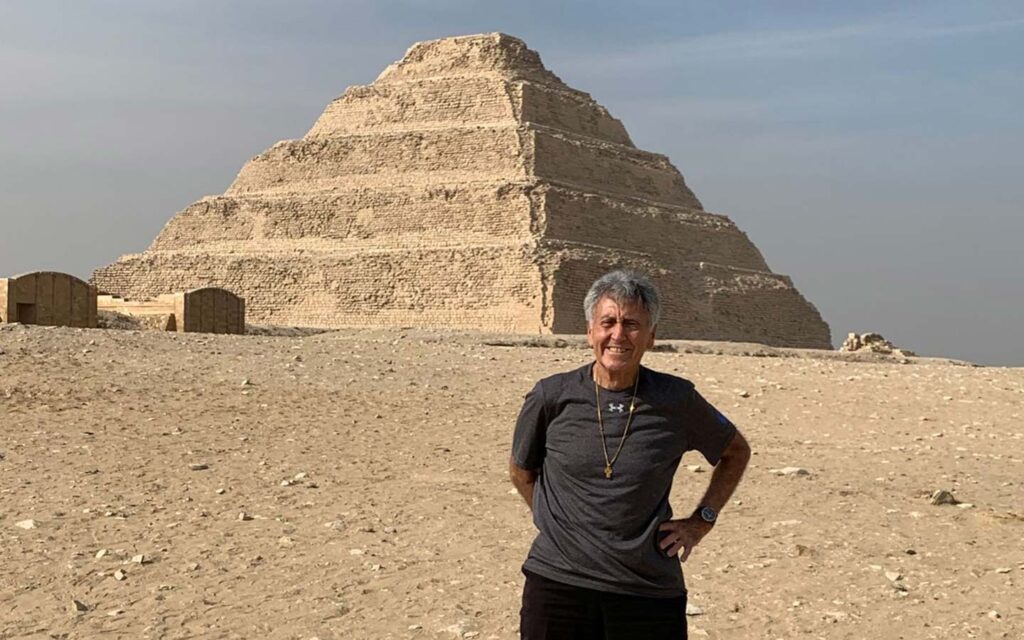 A smiling older man with gray hair standing in front of the step pyramid of Djoser in Egypt on a sunny day, showcasing one of the best sights of his Egypt travel story.