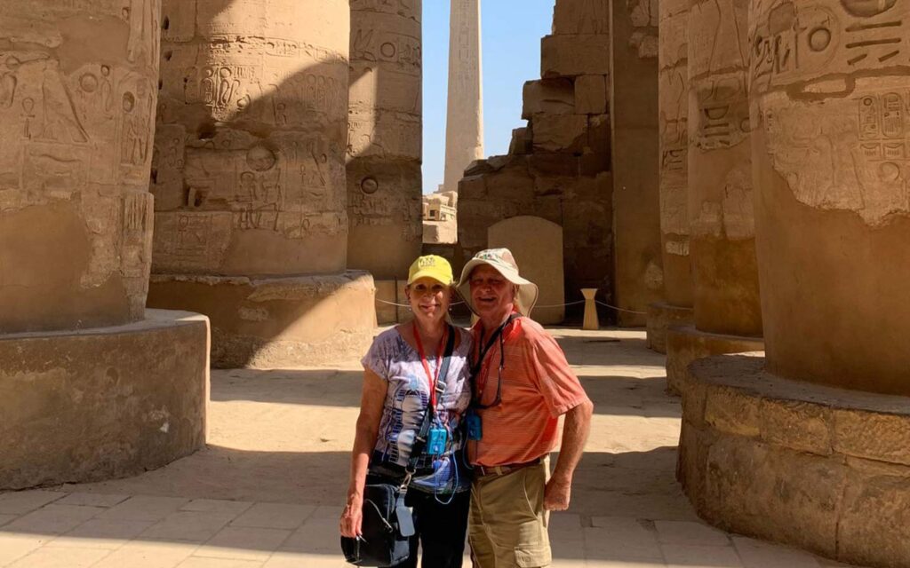 A couple wearing sun hats and sunglasses pose together at the Karnak Temple in Egypt, surrounded by ancient columns with hieroglyphics, capturing their Egypt travel story.