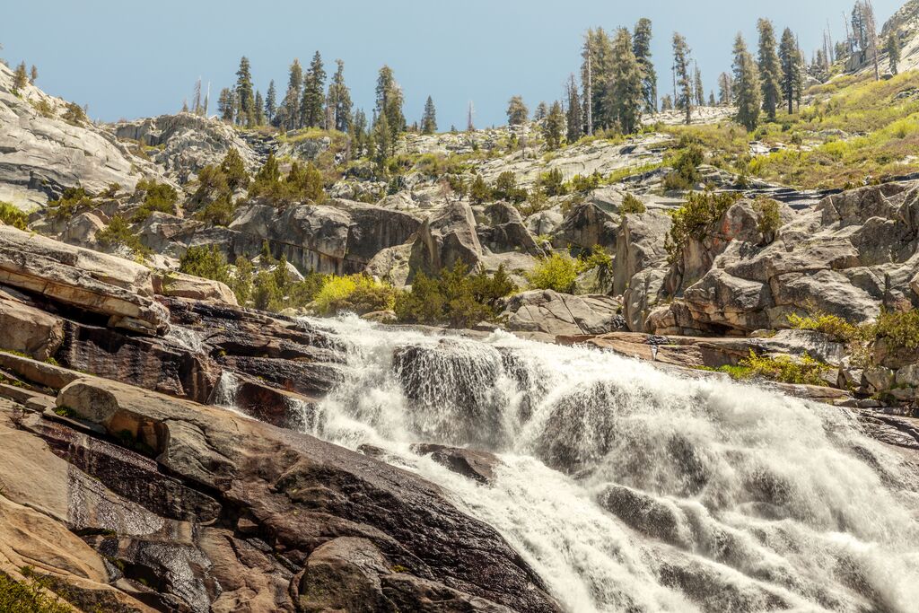 A waterfall cascades over rocky terrain surrounded by pine trees under a clear blue sky, perfect for scenic hikes.