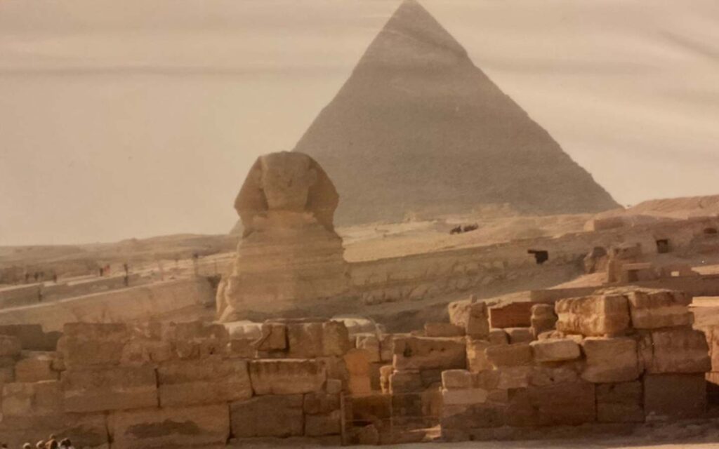 The great sphinx and a pyramid in the background, under a hazy sky, with visible ruins in the foreground invite you to visit Egypt.