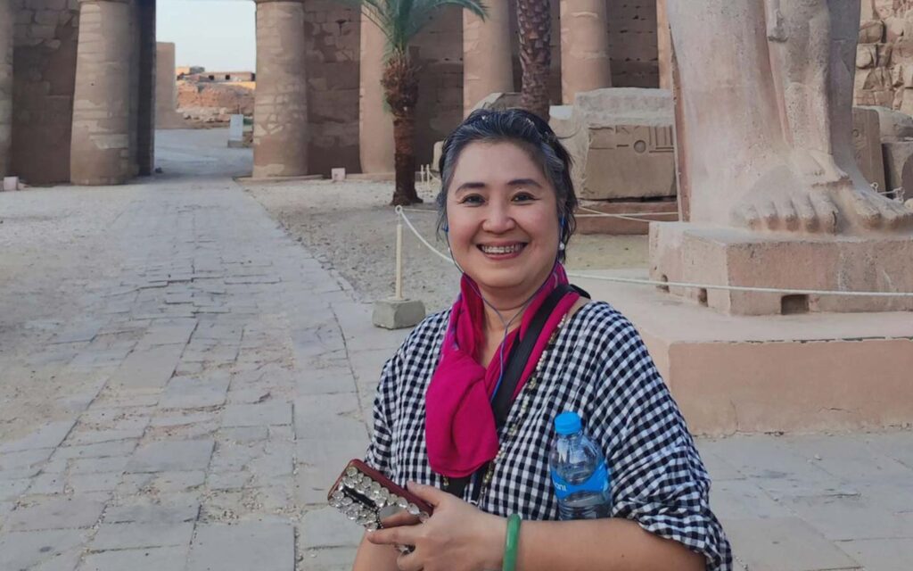 A smiling woman holding a camera stands in front of ancient stone ruins in Egypt, wearing a checkered shirt and a pink scarf. This photo captures the essence of her Egypt travel story.