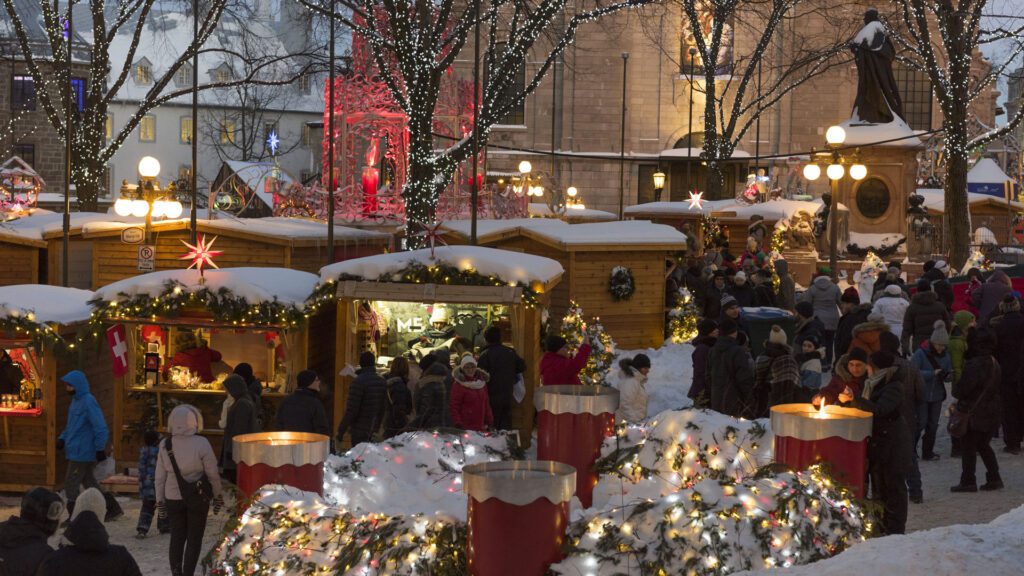 Outdoor christmas market bustling with people shopping at wooden stalls decorated with lights and garlands, exuding christmas charm, surrounded by snow-covered trees and festive decorations.