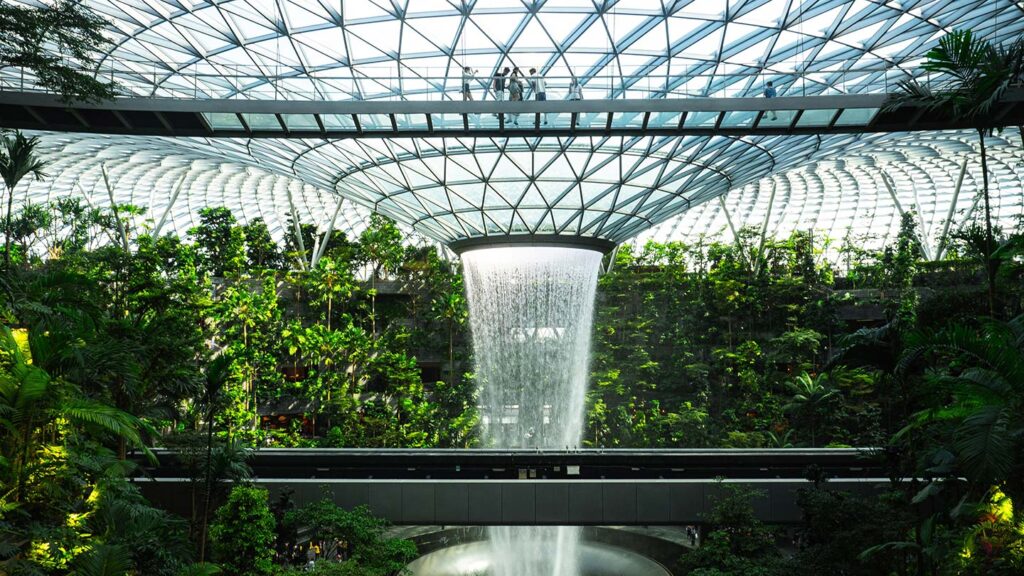 Indoor waterfall cascading down a circular structure, surrounded by lush greenery under a large dome with a geometric glass roof, located in one of the highest cost of living cities.
