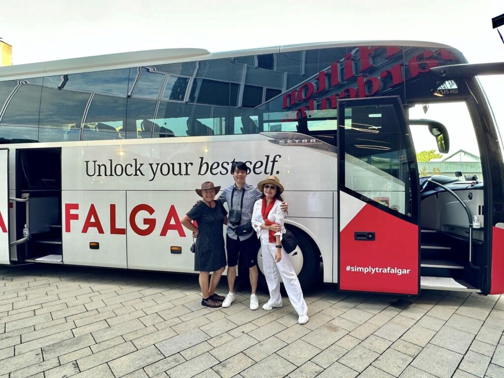 Three people posing in front of a Trafalgar tour bus with the text "unlock your best self" and "#simplytrafalgar" on its side.