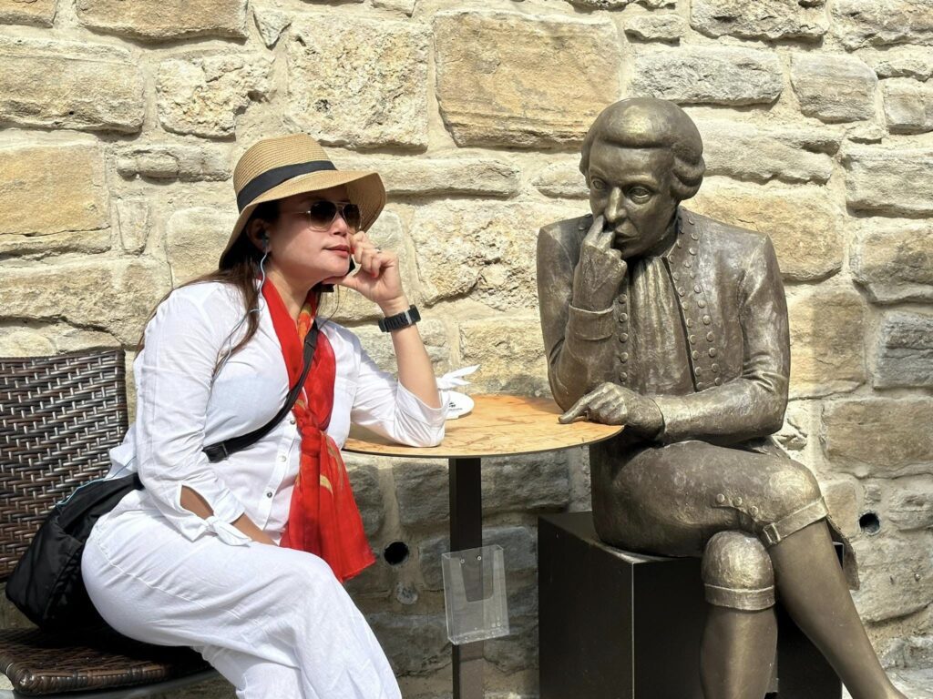 A woman in a white outfit and hat sitting at a Trafalgar tour cafe table, mimicking the pose of a bronze statue of a seated man.