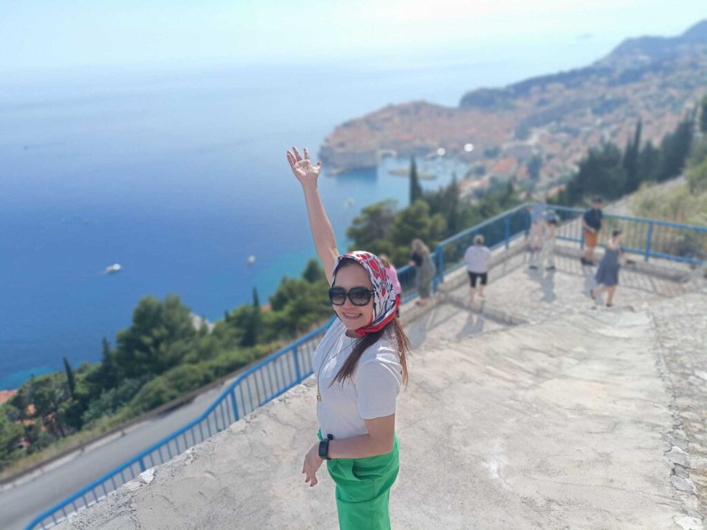 Woman with raised arm smiling at the camera on a coastal overlook with a panoramic view of the sea and a town below under a clear blue sky during her Trafalgar tour.
