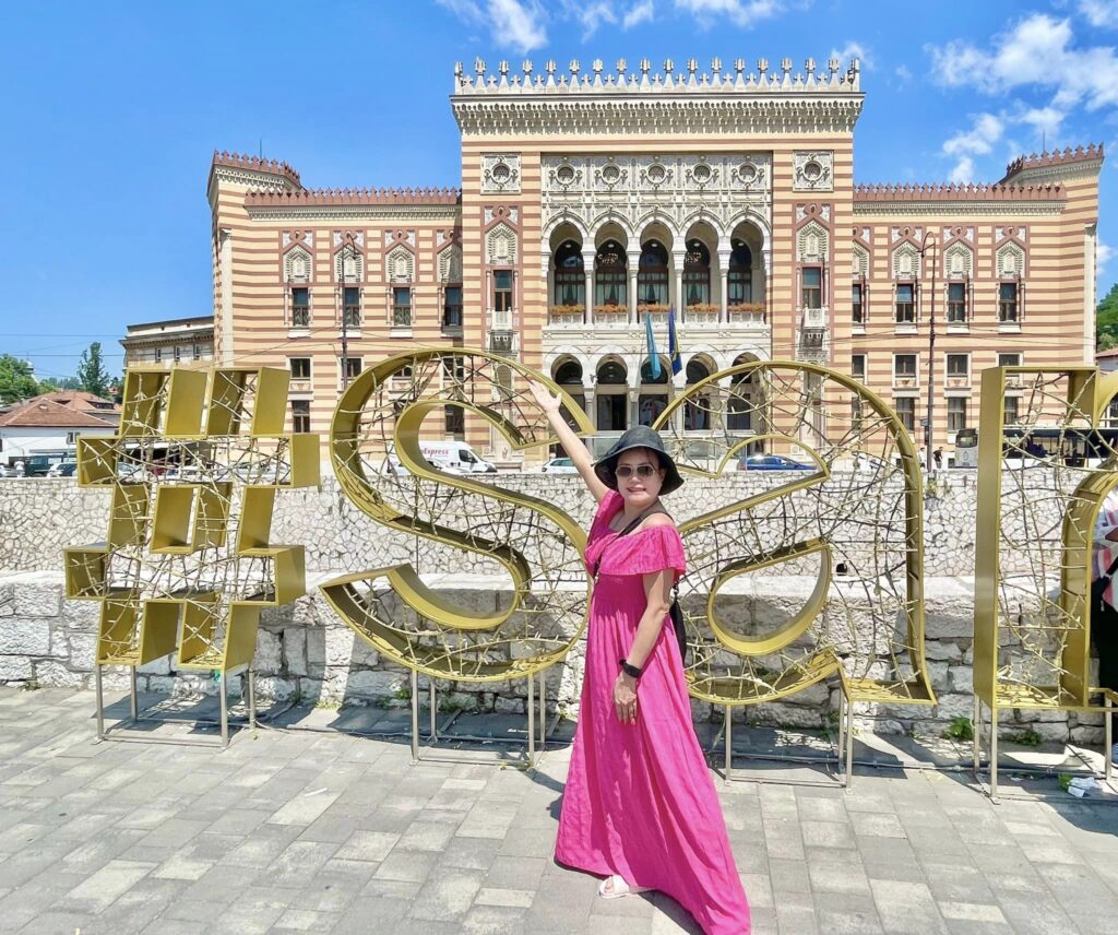 A woman in a pink dress and hat poses in front of a large golden "#stay" sign with an ornate historic building in the background during her Trafalgar tour.