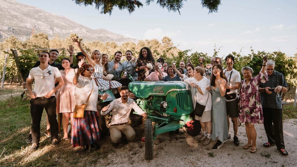 A diverse group of people celebrating joyously around a vintage tractor in a vineyard, with mountains in the background at a destination wedding.