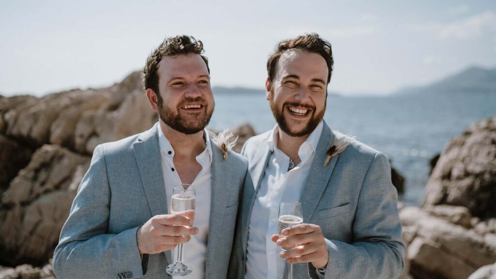 Two men in suits smiling and holding champagne glasses by the seaside on a sunny day, celebrating a spontaneous wedding.