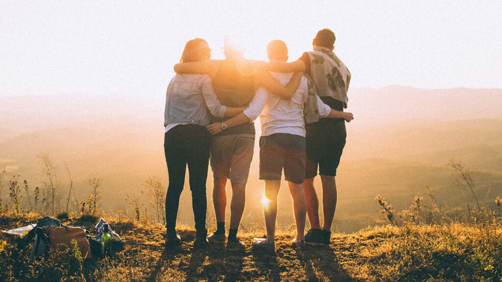 Four friends embracing while watching a sunset over rolling hills, conveying a sense of camaraderie and learning from travel.