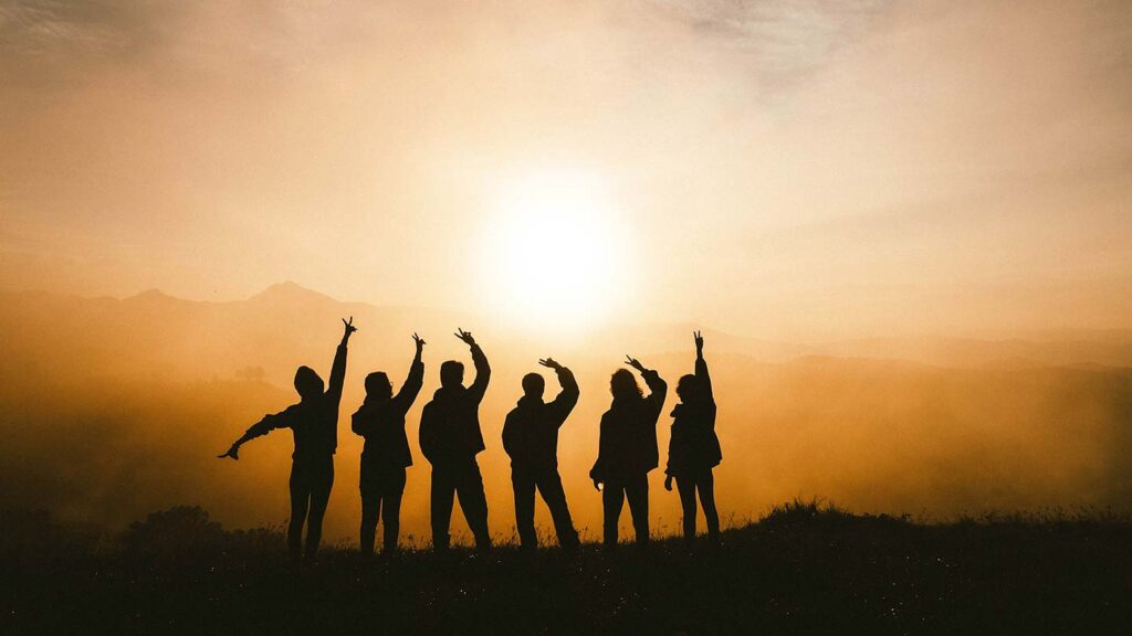 Silhouettes of seven people standing on a hill with arms raised against a sunset and hazy mountains in the background, capturing the essence of how travel heals.