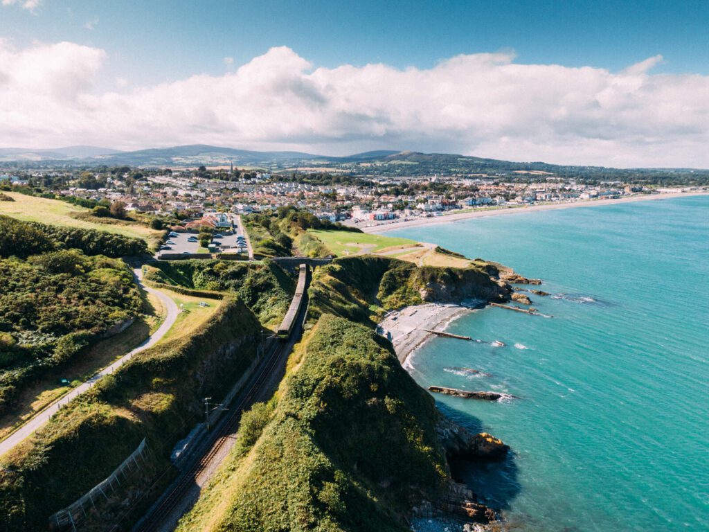 An aerial view of Bray Head Cliff Walk, the water is turquoise, the fields are green, and the most lush of Ireland's landscapes stretch out into the background. 