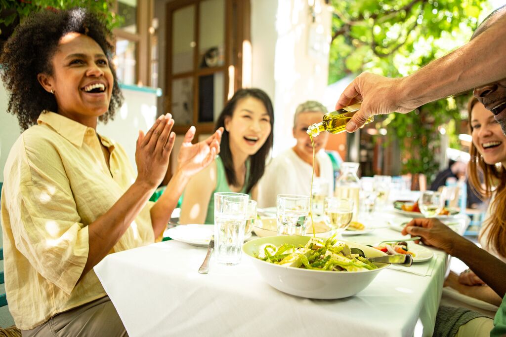 A group of travelers sit at a table, laughing and clapping while a hand tips a bottle of olive oil down onto a bowl of salad.