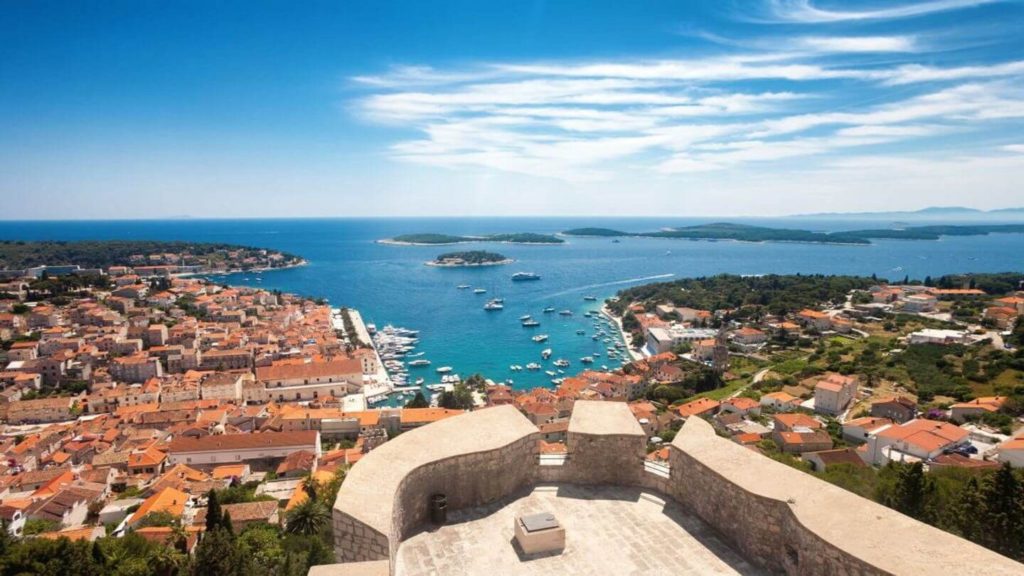 Aerial view of a coastal town with red-roofed buildings and a marina, overlooking a calm blue sea with islands in the distance on a tour with Trafalgar.