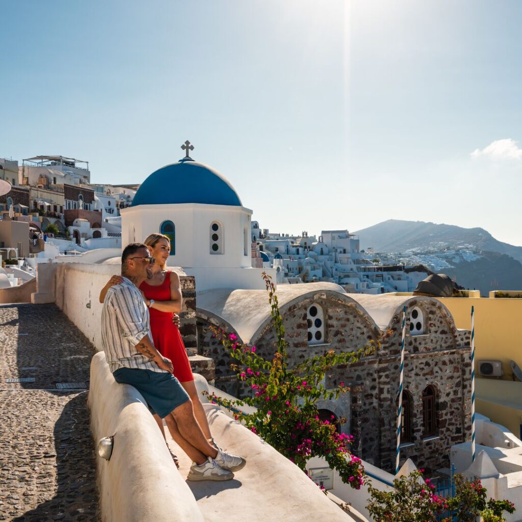 A young couple embrace in Santorini, Greece, with the iconic blue domed buildings behind them.