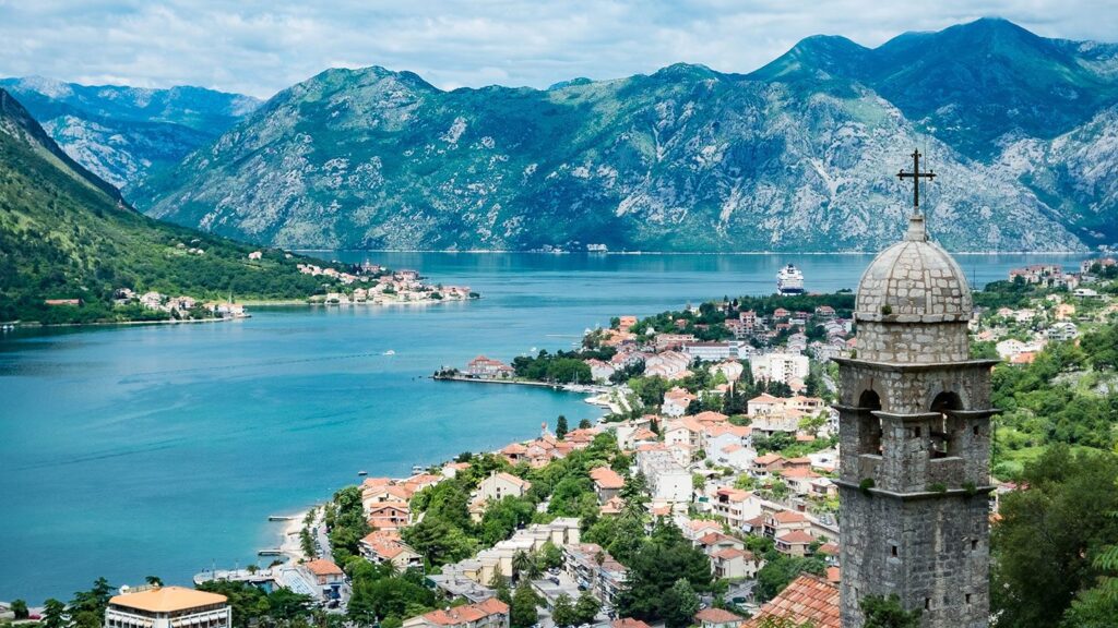 Aerial view of a coastal town with red-roofed buildings, nestled between a tranquil bay and rugged mountains, showcasing a church bell tower in the foreground. Ideal for 2023 tours.