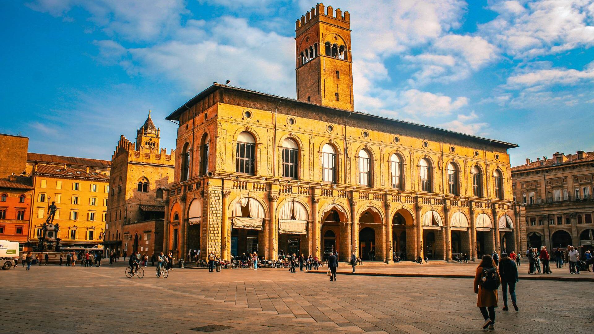 Orange-toned building in Bologna, with people stood all around it. A statue is in the background, with more orange-hued buildings.