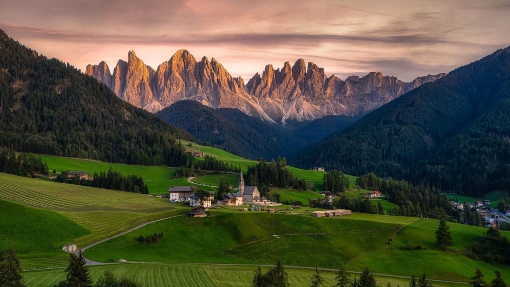 Sunset over a lush valley with a small village, featuring the prominent, jagged mountain peaks of Italy's Dolomites.