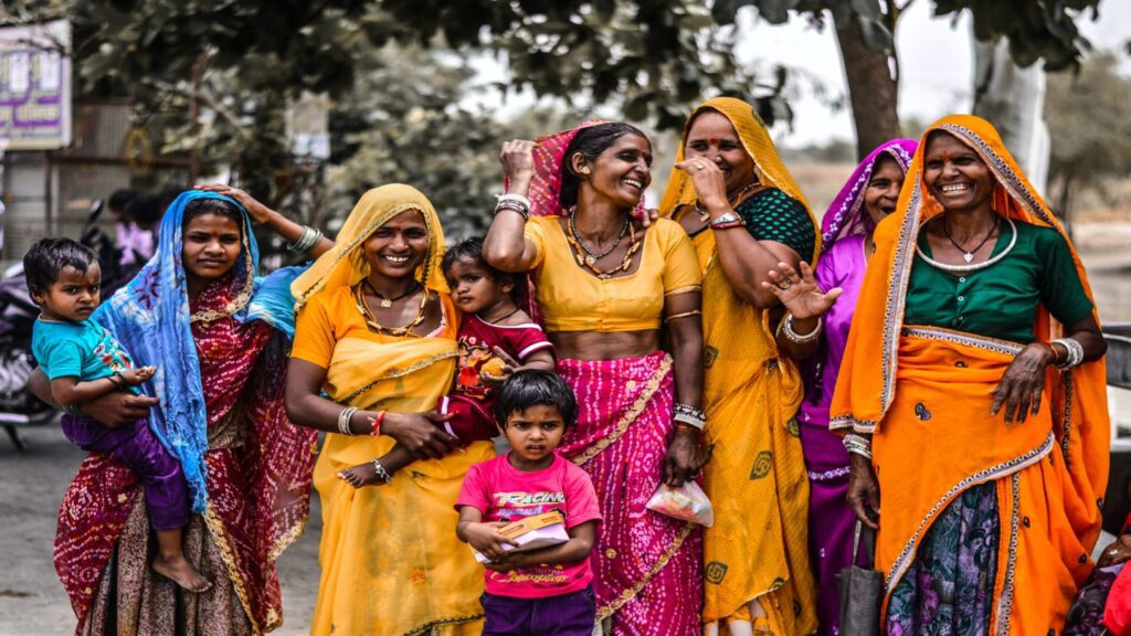 Group of Indian women and children in traditional clothing, smiling and interacting on a rural roadside.